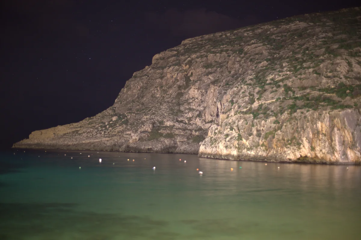 Illuminated cliffs reflected in Xlendi Bay after dark.